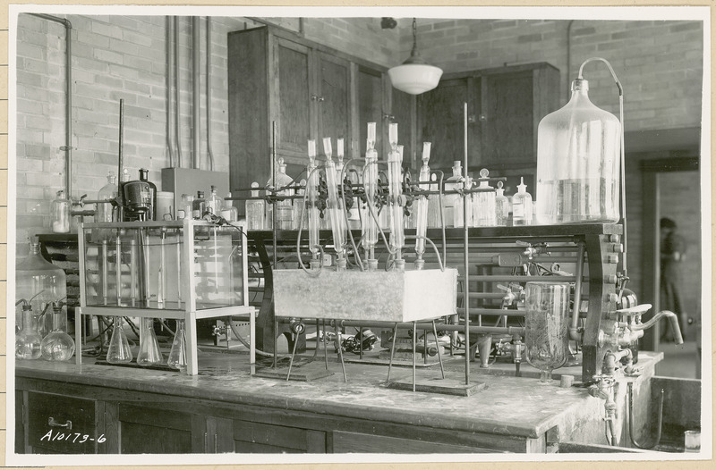 A variety of tubes, flasks, bulbs and other chemical glassware are visible on a laboratory table. A female researcher can be seen in the open doorway.