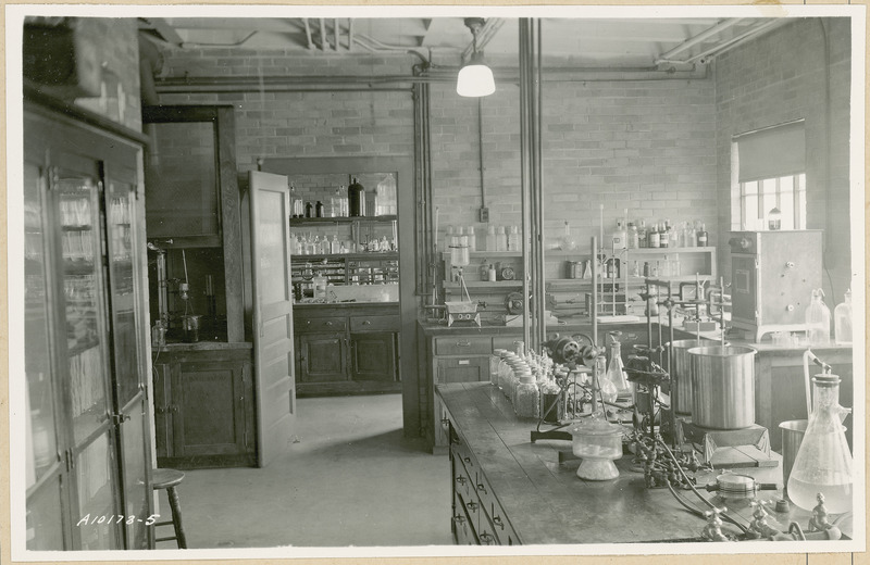 Various glassware and instruments are displayed on tables and counters in this laboratory. A large cabinet with glass doors is on the left. The door at the rear opens into another laboratory.