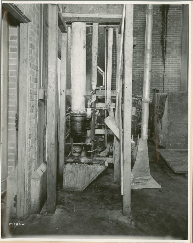 One large and two smaller wrapped vertical pipes are shown in the foreground. A container, wider at the top than the bottom, sits below the larger pipe. A burlap bag is wrapped around another pipe on the right.