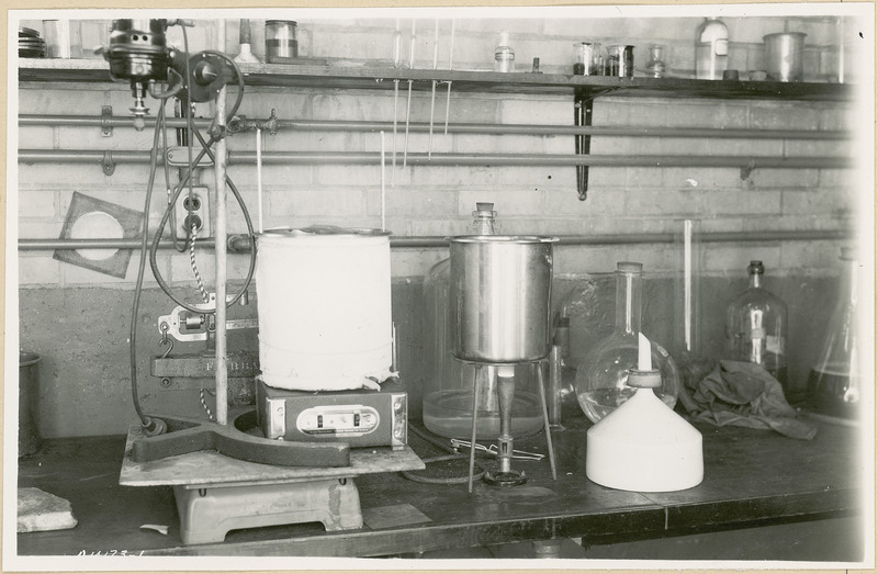 Chemical instruments and glassware are displayed on the table. There is a roll of paper or cloth in front of a Fairbanks scale on the left.