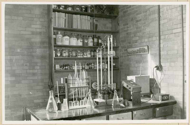 A number of glass instruments are displayed on a laboratory table. The shelves in the background contain glass jars filled with various substances, reference books and scales. The spine of some books on the top shelf have the title, "Paper trade" and dates in the 1930s.