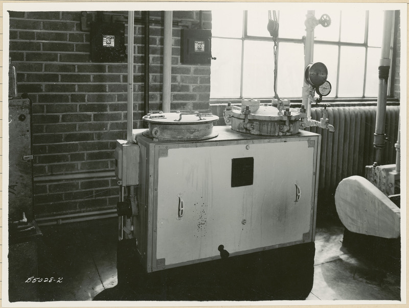 On top of this piece of equipment are two openings with lids. The one on the left has a handle. The one on the right is raised via a pulley system. There are two pressure gauges and a faucet to the right.