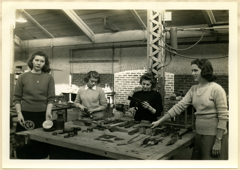 Four cadettes surround a workbench displaying the Guerin (rubber) embossing die, cupping die, dimpling die, drill jigs, templates, soldering jigs, forming blocks and standard hand tools.