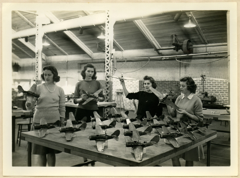 Four cadettes are holding their metal model planes. A group of the unfinished planes at the end of the third period are displayed on the workbench.