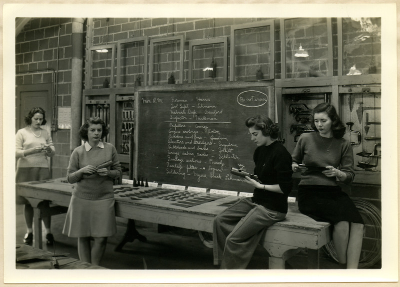 On the blackboard is the organization for one of the laboratory periods. The cut and formed parts are on the table for inspection before assembling begins. Two cadettes are standing to the left of the blackboard and two are seated on a table to the right of the blackboard. Each cadette is holding a part. There are tools hanging in cases on both sides of the blackboard.