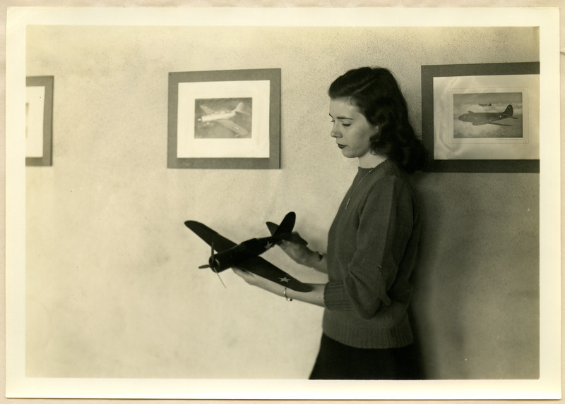 Jane Ragsdale is holding one of the completed model airplanes. Photographs of aircraft are hanging on the wall behind her.