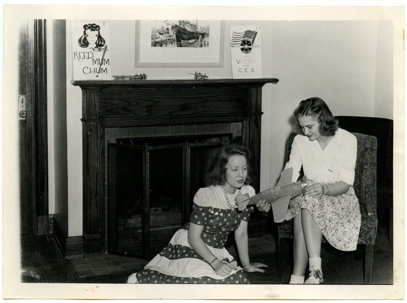 Betty Cue, a stenographer at the Memorial Union, is seated showing a Curtiss P-40 Warhawk model plane to a cadette who is sitting on the floor in front of a fireplace. The plane has a wood framework with a paper exterior with a wind up, rubber-band driven propeller. A poster on the left side of the mantel reads, "Keep Mum Chum" and the one on the right reads, "Curtiss for Victory with C.E.S.".