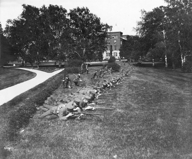 A cadet battle drill with weapons. Officers are carrying swords. An officer mounted on a horse can be seen in the distance in front of Buchanan Hall, which is partially visible through the trees.