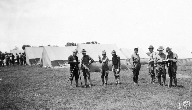 Activities at training camp. In the foreground in front of a tent encampment a squad of eight men are engaged in making maps. On the image left a group of men are in line to move out of the group of tents.