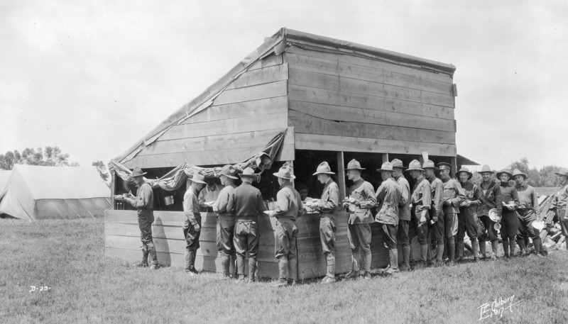 A military squad at camp lined up at a mess hut. Seventeen men are in the line.