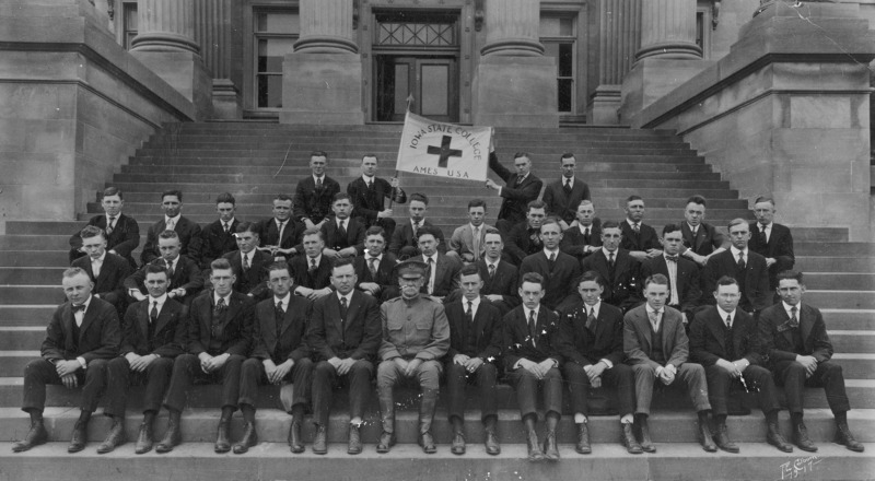 Male students formally sitting on the front steps of Beardshear Hall with a man in military uniform. In the back row two men hold a banner with the text "Iowa State College, Ames, USA" surrounding a cross.