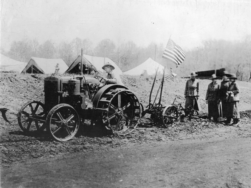 "American girls learning to use tractors in order to increase the Country's food supply at the National Service School, Inc., of the Women's Naval Service, Inc." The tractor and the plow being pulled have all metal wheels without tires. An American flag is mounted on one of the control levers of the plow.