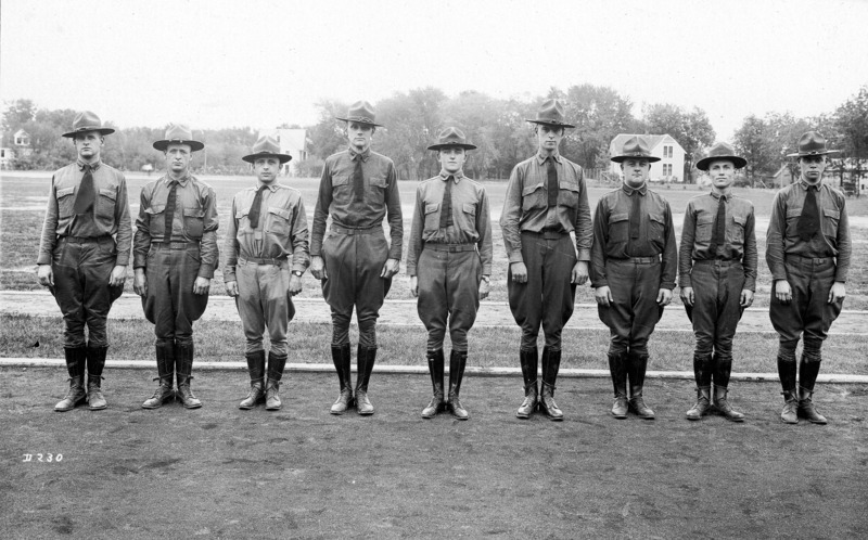 Nine officers in military uniform standing at attention in pose for a photograph. They are standing on an athletic track with residential housing in the distance across an open field.