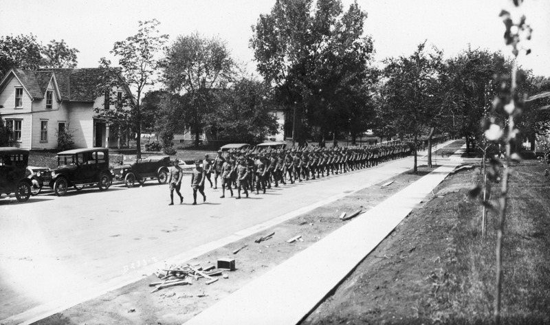 A street scene in which a large training detachment of U.S. soldiers in uniform is marching in formation with rifles on shoulders. Cars are parked on one side of the street. In the foreground there is debris that suggests a sidewalk installation had just recently reached completion. Residential housing is in the background.