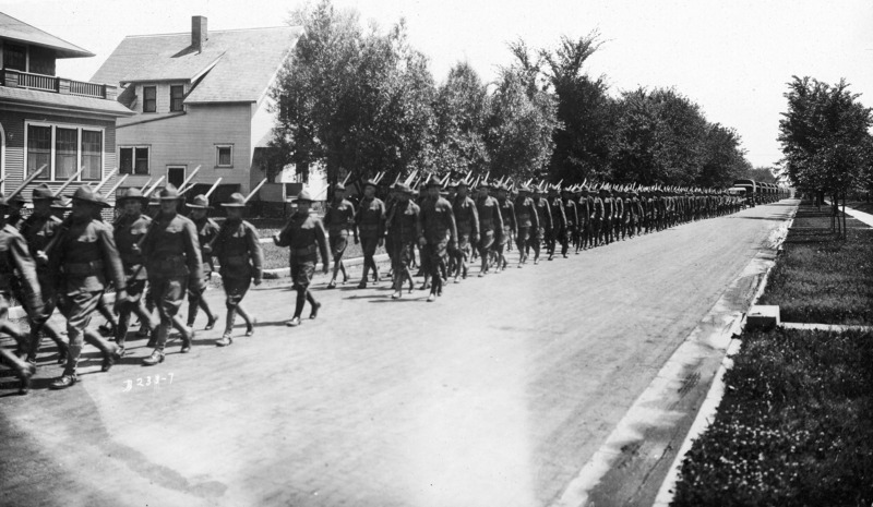 A large U.S. Army detachment in uniform marching on a city street in formation with rifles on shoulders. Following the soldiers is a convoy of military transport trucks. Residential housing is in the background.