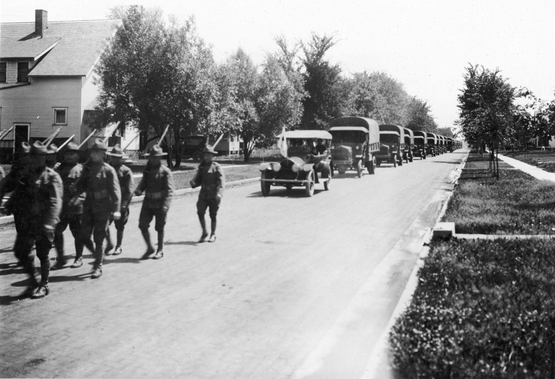 The rear of a marching U.S. Army detachment and the beginning of the vehicle convoy following the marching soldiers. The military transport vehicles are preceded by a civilian vehicle with a banner mounted on the hood. Residential housing is in the background.