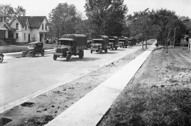 A military vehicle convoy on a city street. Civilian vehicles are parked on one side of the street. Residential housing is in the background.