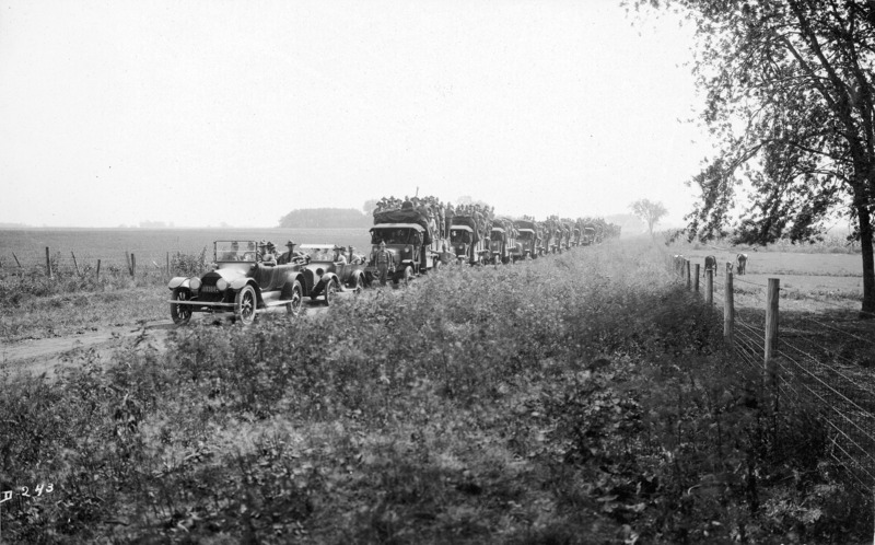 A U.S. military convoy on a dirt road to Boone, Iowa. The truck convoy is lead by two civilian vehicles with the tops down and five men in each vehicle. The transport trucks have canvas tops rolled back with men standing in the truck in a pose for the photograph. One officer is standing at attention at the front of the lead truck; another officer is leaning on the truck. Two cows are grazing in a pasture on the image right.