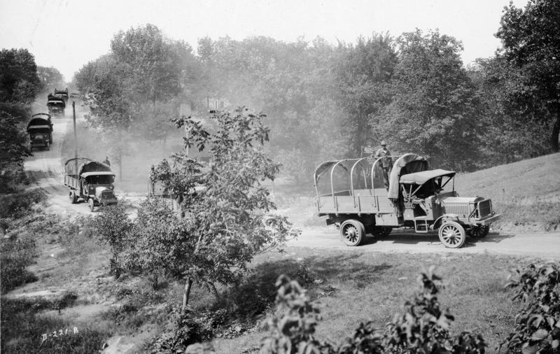 A U.S. military training convoy on a dirt road:"liberty trucks in a train across the country." The transport trucks have canvas tops rolled back with one man standing in each truck in a pose for the photograph.