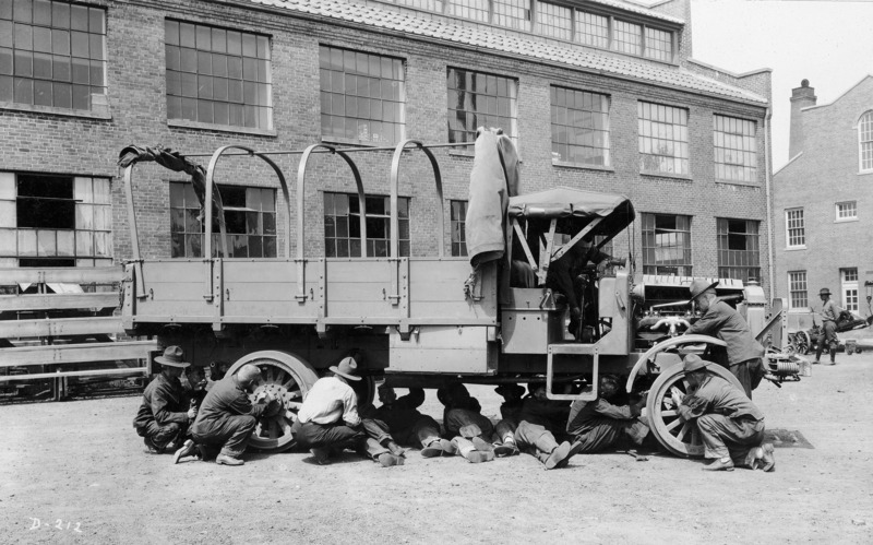 A U.S. army training exercise. Twelve men are working on a military transport truck. A brick industrial building is in the background.