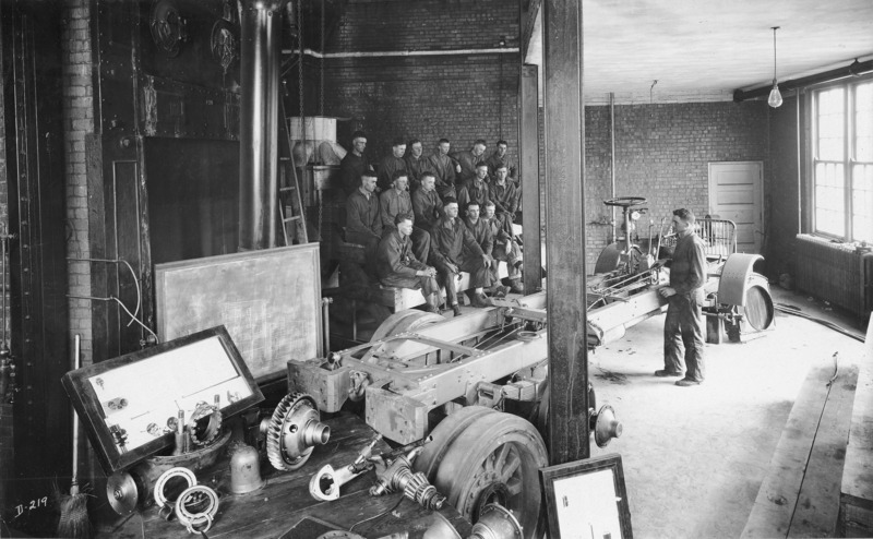 A U.S. army training course. Fifteen men are sitting on a classroom bleacher as a man standing by a truck undercarriage lectures.