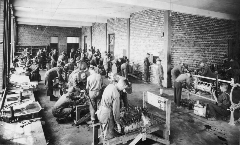 A U.S. army training course. A detachment of men are engaged in various aspects of vehicle engine repair. Military officers are depicted as observers.