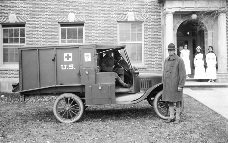 A U.S. army ambulance with two uniformed men, one in the driving seat and one standing beside the vehicle. The Student Services Building is in the background, at the front door of which three uniformed nurses are standing.