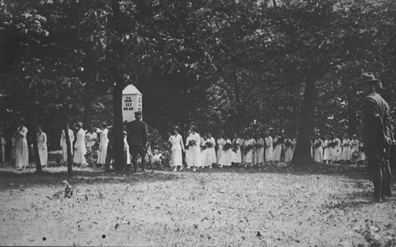 A military honorary event in the ISC campus cemetery. A file of approximately thirty women dressed in white and carrying flowers is passing a structure on which is lettered "To our 117 dead." An older officer with a cane, back to the camera, faces the oncoming women. On the right side of the image a line of uniformed men stand at parade rest.