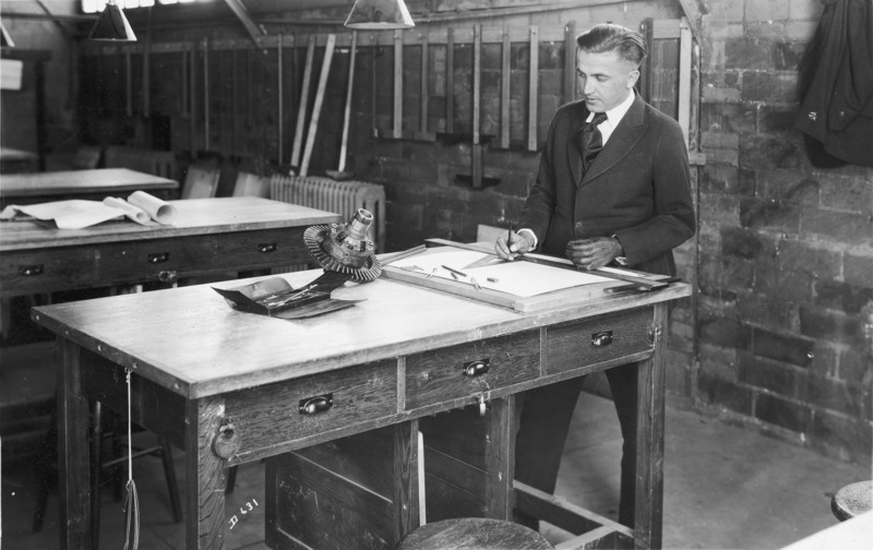 A young man in a suit with drafting equipment. He is posed looking at a gear as if he is about to begin an engineering drawing of the object. Drawing tables fill the room; on the back wall hang a large number of t-squares.