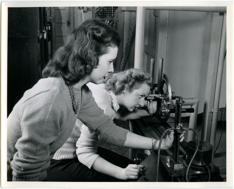 One of the cadettes is using an optical telescope system, possibly to view the test model inside the wind tunnel. The cadette on the right is monitoring another instrument.