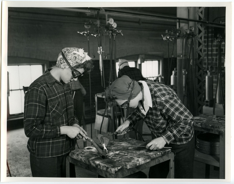 Two cadettes are welding metal parts together. They are wearing slacks and eye goggles and their hair is wrapped in a turban.