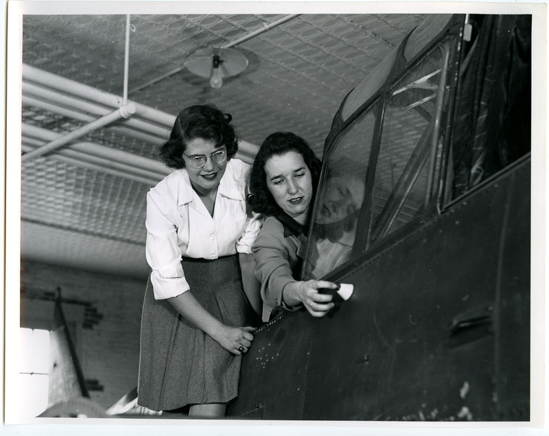 A cadettes is touching the top of a small cone-shaped part on the side of a small airplane in the Aeronautics Laboratory. This plane, sans wings, was used for instructional purposes. Another cadette is by her side also looking at the part.