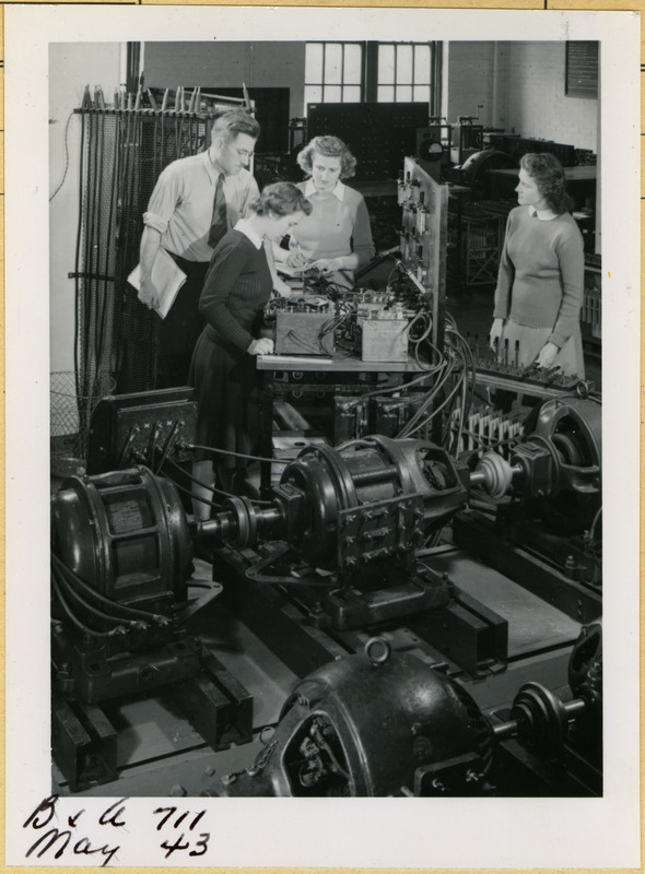 Three cadettes are listening to instructions from their Professor Benjamin Willis in the Electrical Engineering Power Laboratory. One cadette is focused on a piece of equipment while another is taking notes. The third cadette looks on from the right. Two rows of electric motors are in the foreground.