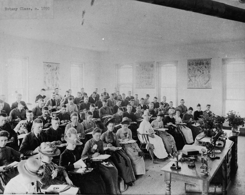 The Botany class is sitting in a North Hall classroom. The women students are sitting near the front of the room. Microscopes , books, and some flowers are in vases on the instructor's desk.