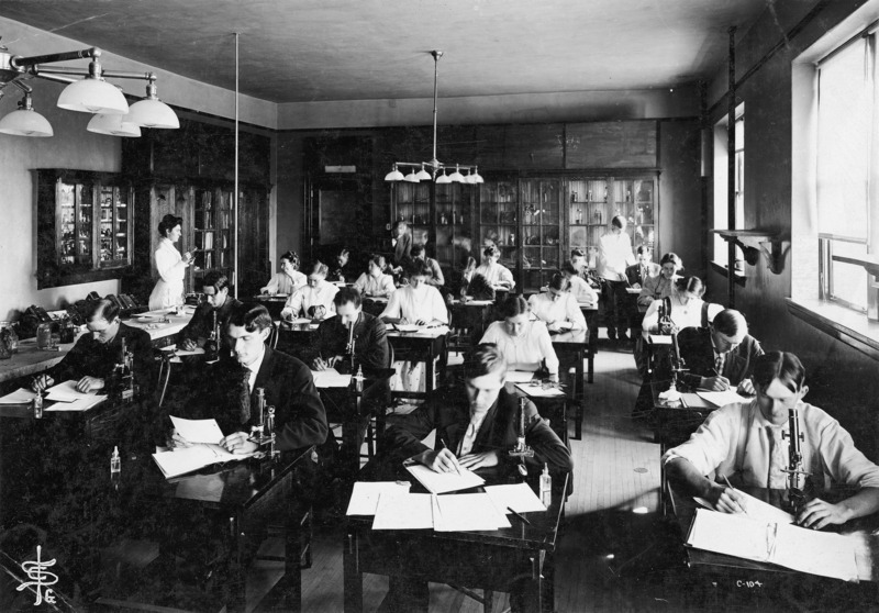 Students are writing at their desks in the Botany Lab in Central Building (Beardshear Hall). Some students have microscopes on their desks. Dr. Pammel, wearing a suit, is assisting a student at the back of the lab. A man wearing a white jacket is assisting another student. Estella Fogel (Buchanan) may be the woman standing at the left.