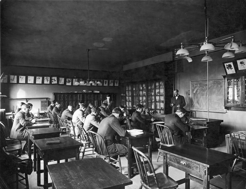 Dr. Pammel is lecturing a Botany class studying weeds in the Botany Lab located in the Central Building (Beardshear Hall). Some of the students are using microscopes. Botanical specimens are framed and hanging on the walls.