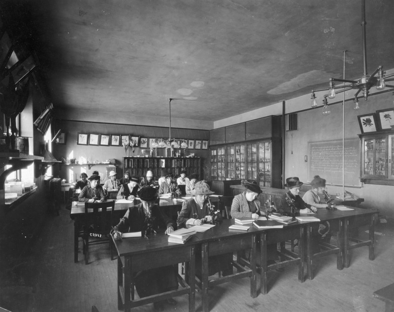 A class of all female students is working in the Botany Lab in Central Building (Beardshear Hall) perhaps completing an assignment written on the blackboard. Books and microscopes are on their desks.