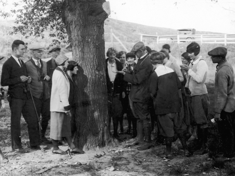 A professor is showing students something in the palm of his hand during a nature study class. The group is gathered around a tree and some of the students have binoculars hanging around their neck. A 1920's car is parked beyond the fence.