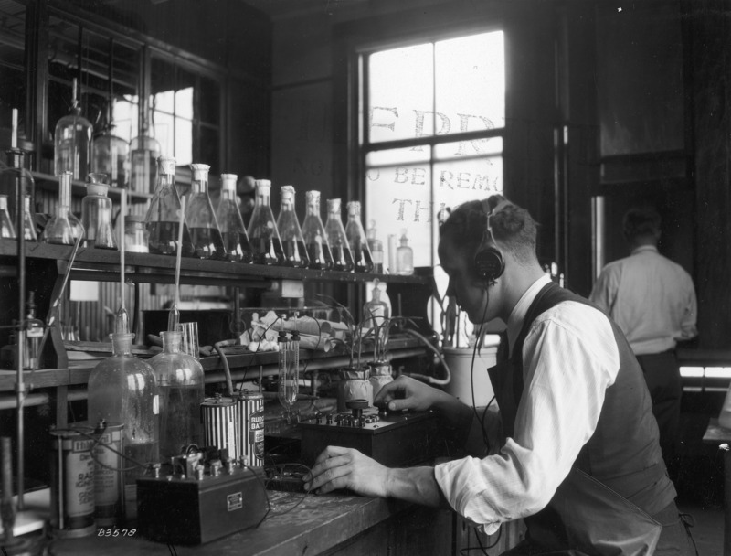 A researcher, wearing a headset, is conducting an experiment on two plants in the Botany Lab. Another man is working in the background.