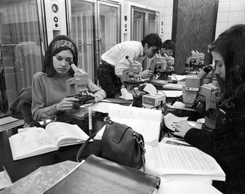 Students are shown examining microscope slides in a laboratory in Bessey Hall. Both monocular and binocular microscopes are in use. Behind the students are climate and light controlled chambers used for experimental purposes.