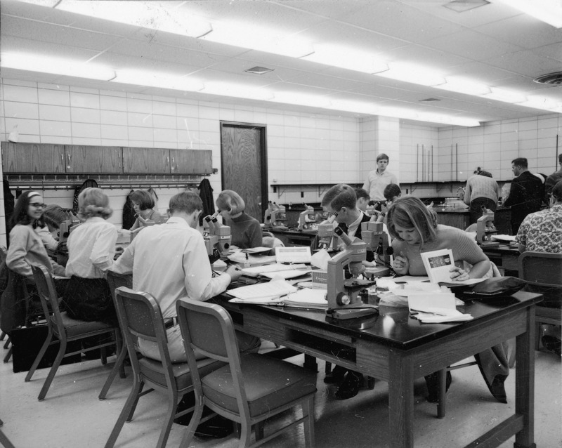 Botany students are sitting at tables in a Bessey Hall classroom. There are microscopes on the tables and some students are working at a lab table at the back of the room.