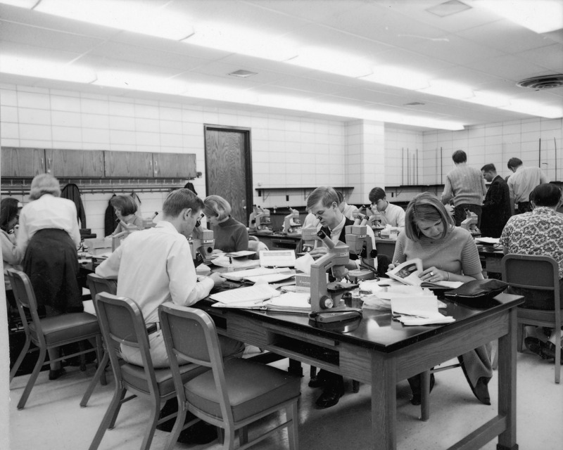 Botany students in a large laboratory room in Bessey Hall. They are shown examining microscope slides using either monocular or binocular microscopes. On the study tables are books, notebooks, and microscopy supplies, including kimwipes and dropper bottles for slide reagents.