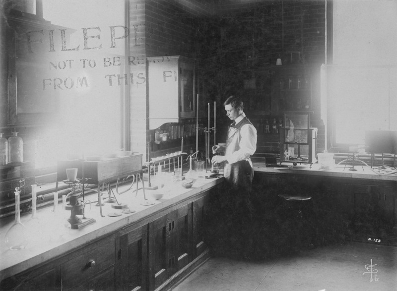 A male student at the bench in a chemistry laboratory. A large number of equipment pieces (funnels, flasks, beakers, burets) are set out on the bench. The date of the photograph according to a note on the reverse is October 25, 1906.