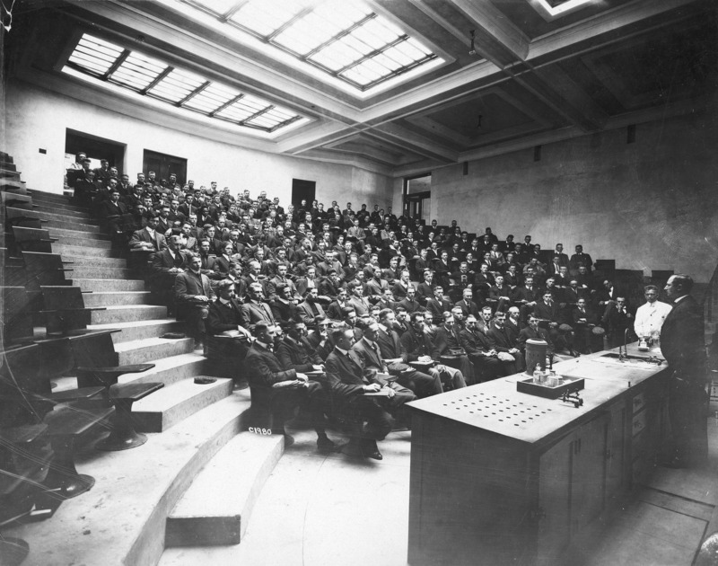 This photograph shows a full class in the chemistry lecture auditorium. A lecturer and an assistant are at the front of the class on the right of the image. The class appears to be all males. The photograph emphasizes the original sky light ceiling in the stadium design hall.