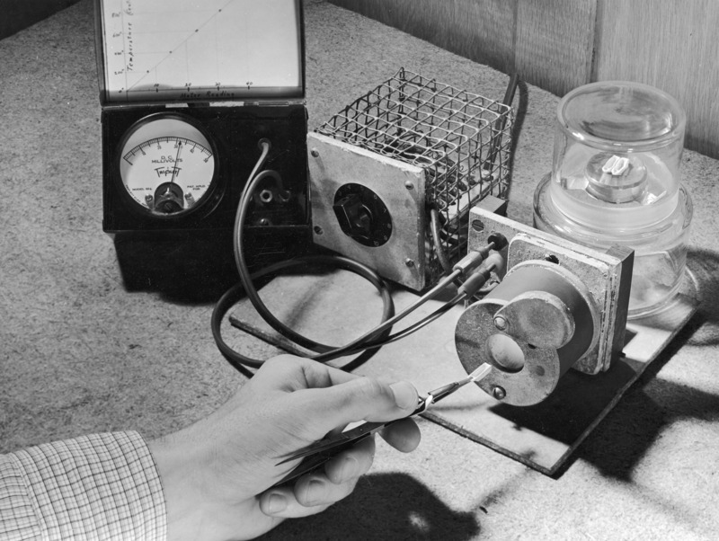 An experiment in micro chemistry, Chemistry 102, in the spring of 1939. The close up photograph shows a hand holding tweezers which in turn hold a small chemical boat as it is about to be inserted into a small electrified chamber. A voltage meter and a home-made rheostatic device are wired to the small chamber.