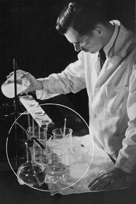 A man in a lab coat pouring a liquid into a funnel, below which are several glass beakers. A volumetric flask is in the foreground. The glassware is circled in white as if for publication purposes.