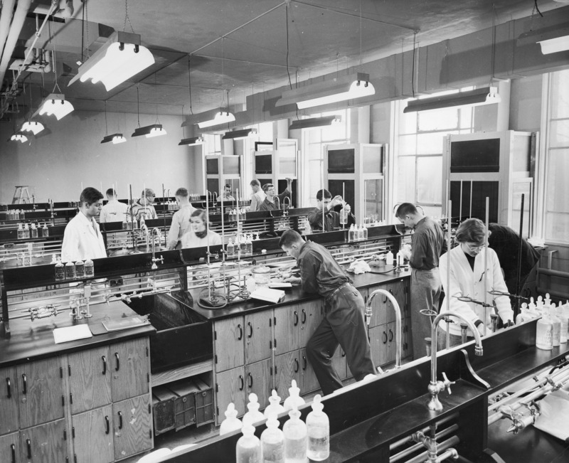 A chemistry laboratory session. Twelve young men and women are working at their individual work stations. A row of fume hoods is against the back wall between large windows. Common laboratory apparatus can be seen throughout the laboratory.