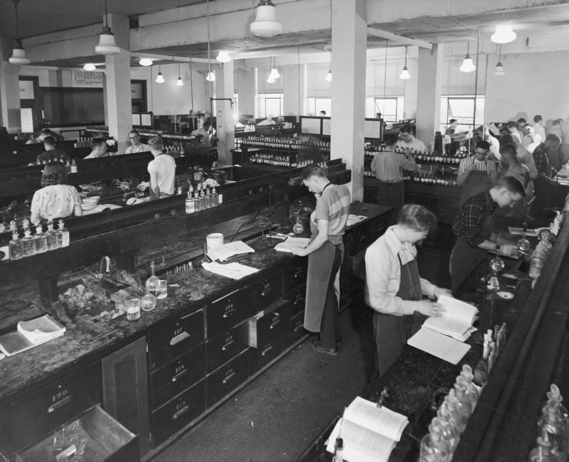 A freshman chemistry laboratory work session. Young men and women are working at their individual work stations. The lab is divided by a central aisle into two sets of work stations. A reagent supply cabinet runs down the length of the central aisle.