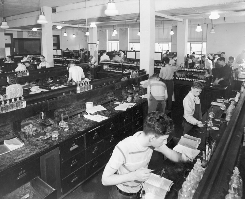 A freshman chemistry laboratory work session. Young men and women are working at their individual work stations. The lab is divided by a central aisle into two sets of work stations. A reagent supply cabinet runs down the length of the central aisle. This photograph is very nearly identical to no. 1052-2-4 except that there are two different students at the work counter in the foreground.