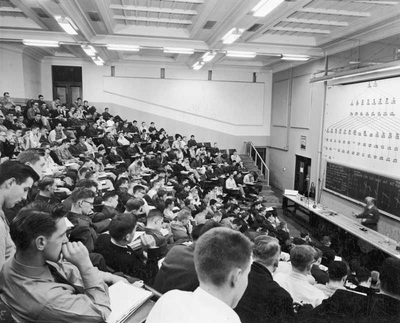 A chemistry lecture class being conducted in the stadium lecture hall within the Chemistry Building (now Gilman Hall). A lecturer stands behind a long demonstration counter at the front of the class. A large periodic chart is on the wall above the blackboard which is behind the lecturer. This photographic view is from the rear audience right looking toward the lecturer.